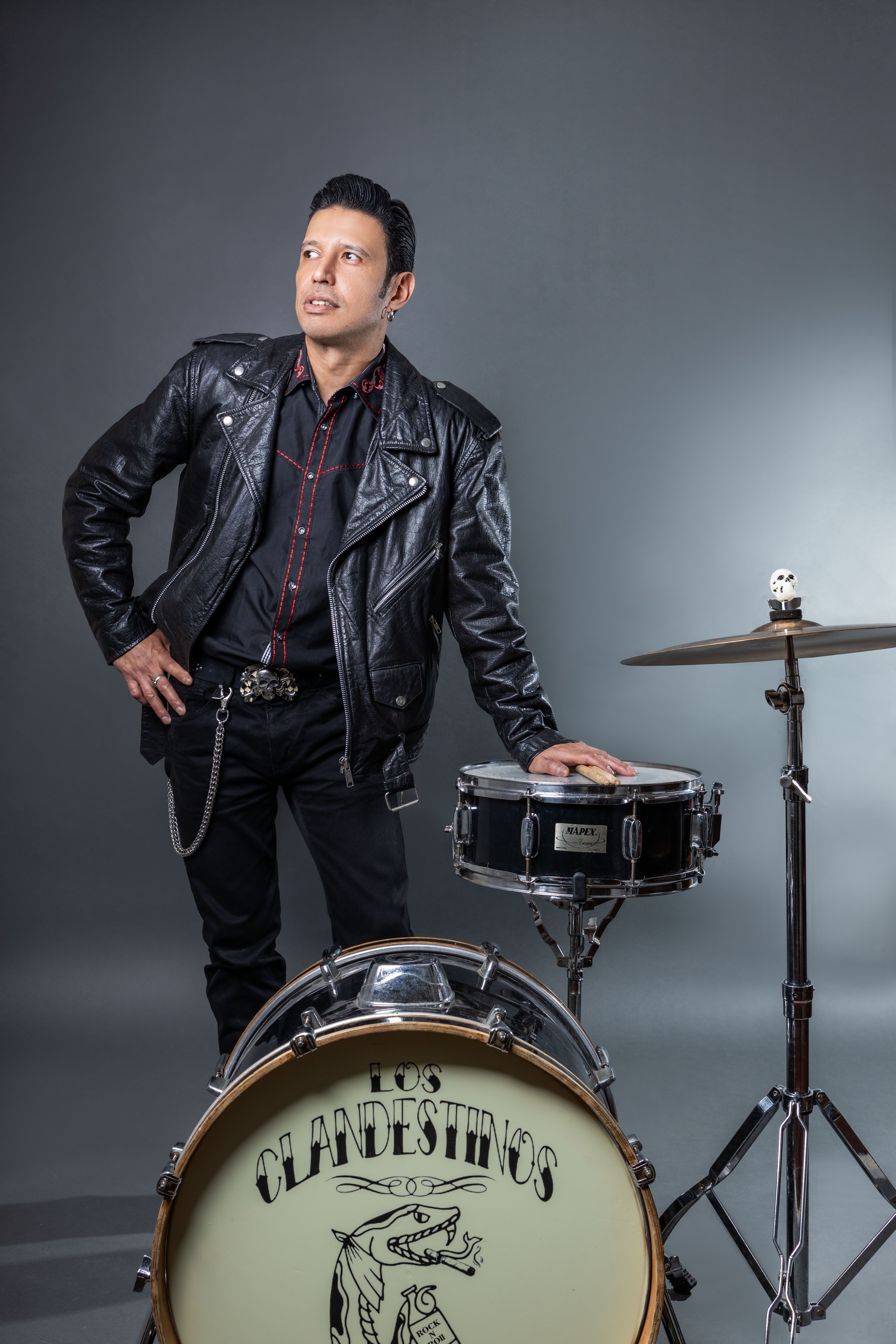 Rock musician in leather jacket poses with drum kit in studio setting.