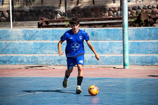 A young male soccer player in a blue uniform dribbling during a street soccer game outdoors.
