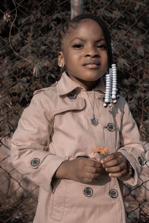 Free A young girl in a stylish trench coat holding a toy, standing outdoors against a chain-link fence. Stock Photo