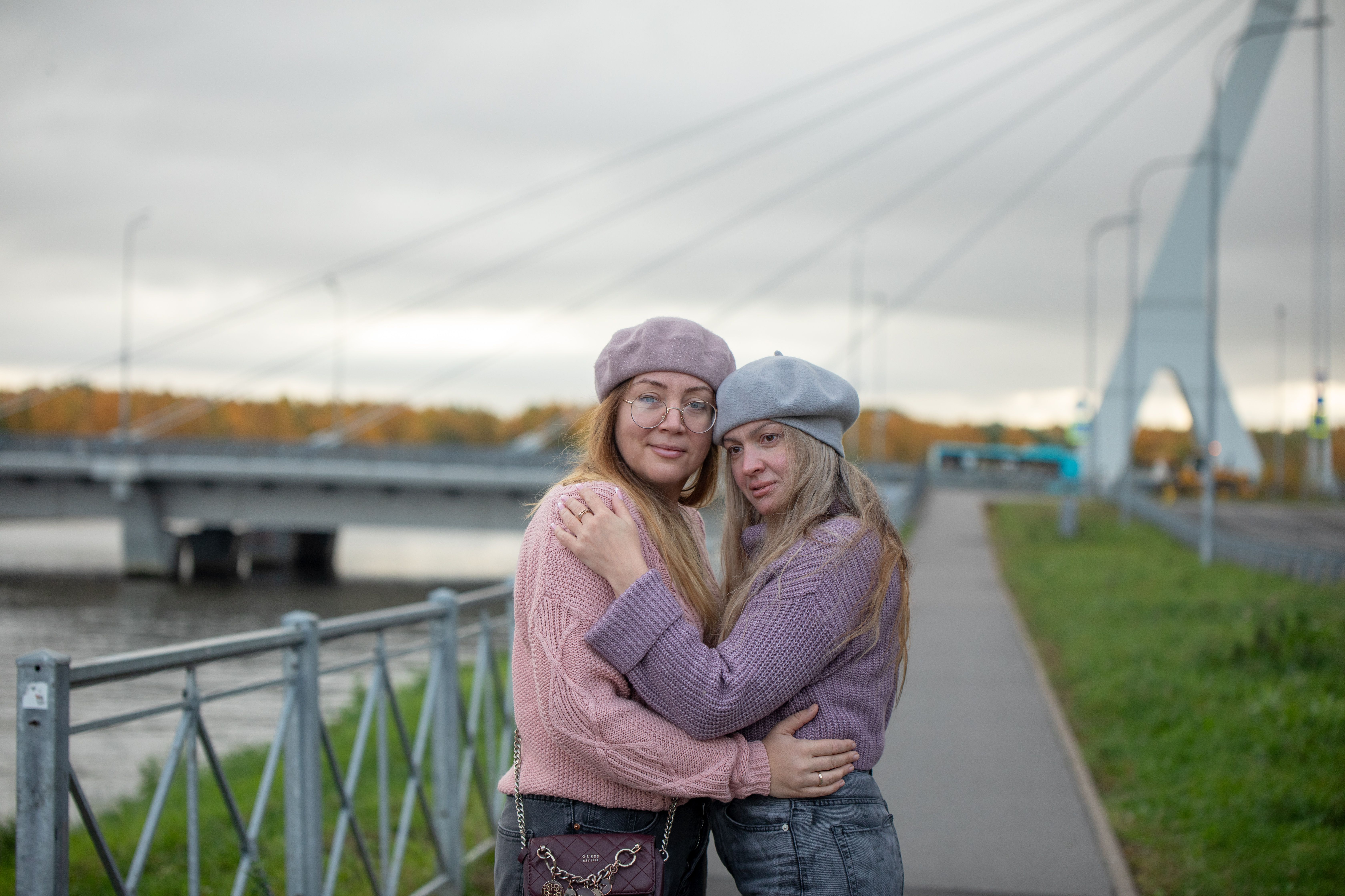 Two women embracing on a bridge walkway in fall clothing, serene outdoor setting.