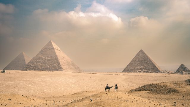 Egypt, Explore the majestic Pyramids of Giza under a sunny sky with distant travelers on camels.