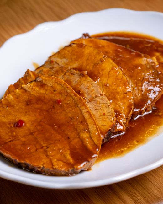 Thinly sliced top round roast fanned across a wood cutting board with a small pitcher of pan jus and fresh rosemary sprigs alongside.