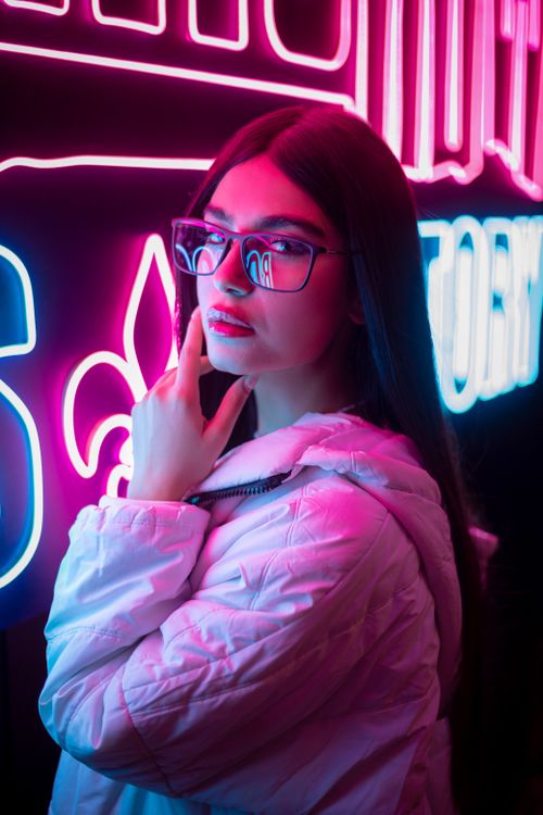 Free Stylish woman in glasses and jacket posing by neon signs, featuring vibrant pink and blue glow. Stock Photo