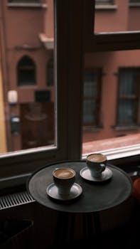 Two coffee cups on a table by a window overlooking an urban scene, creating a cozy atmosphere.