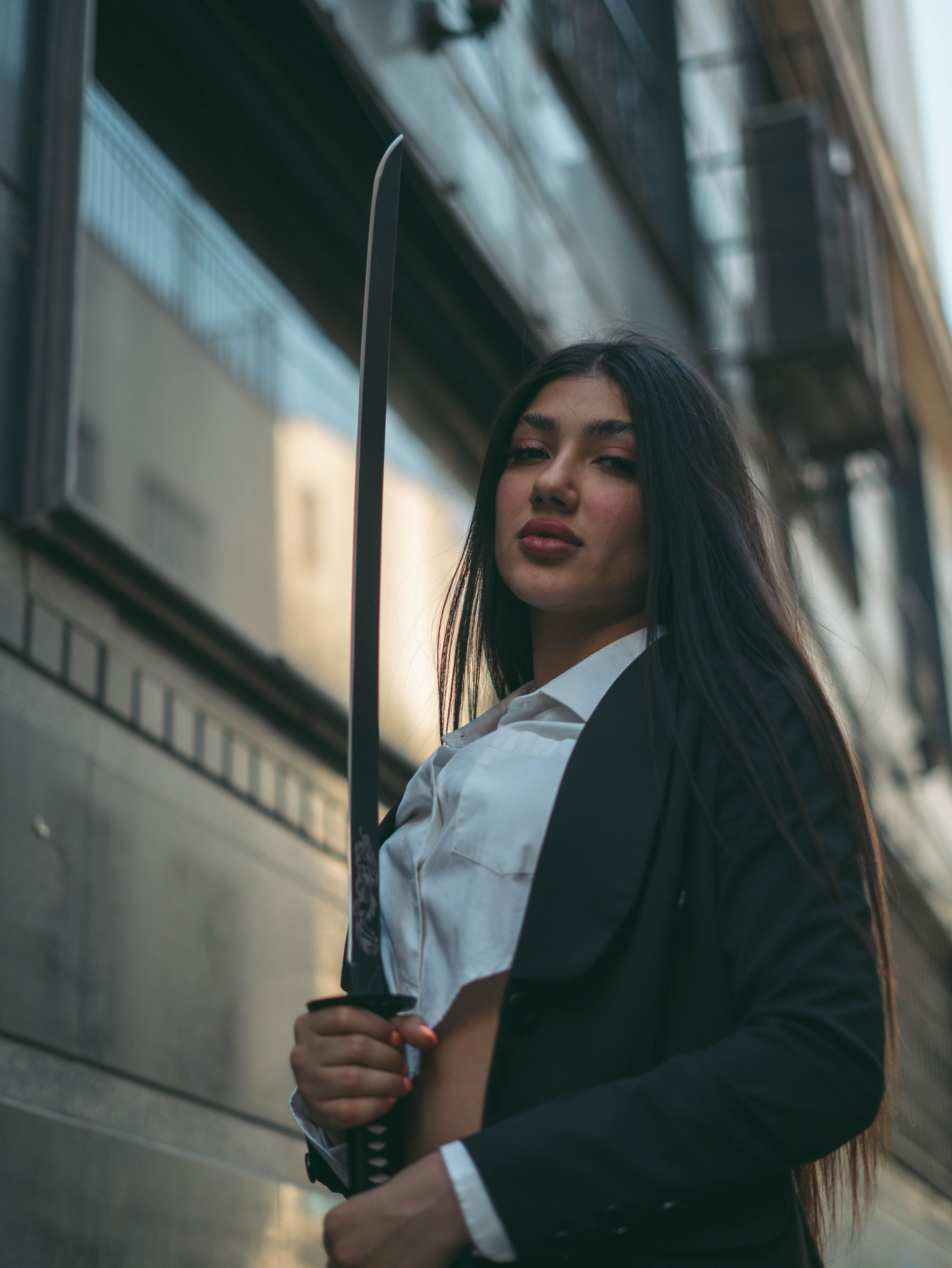 A confident woman in business attire holding a katana sword in an urban alleyway.