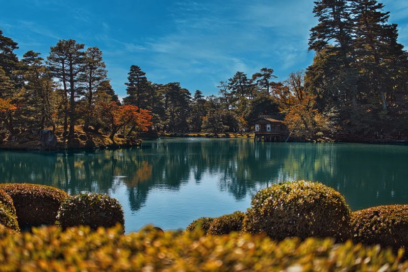 Kenroku-en garden autumn view — tranquil pond reflecting autumn foliage and topiary shrubs, the refined landscape of one of Japan's three great Edo-period gardens