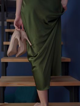 Sophisticated woman in green dress holding high heels while stepping down wooden stairs indoors.