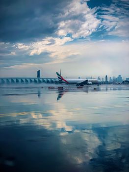 Emirates airplane on wet runway reflecting cloudy sky with city skyline.
