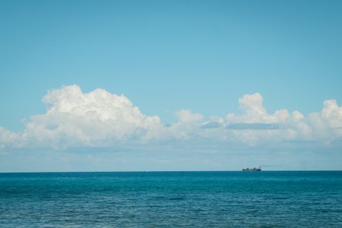 Free Tranquil ocean view with distant ship under a bright blue sky and fluffy clouds. Perfect for serene travel themes. Stock Photo
