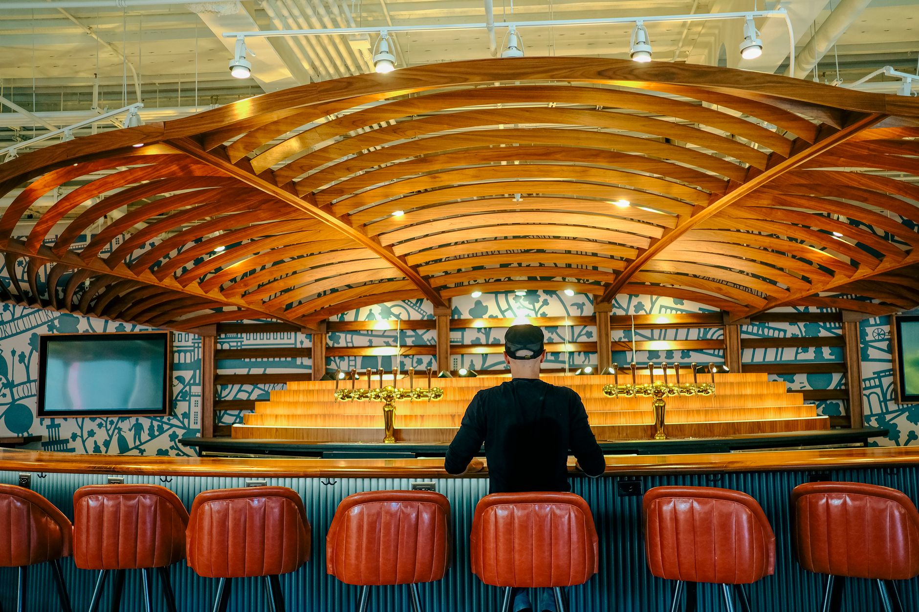 A man sits alone at a stylish bar with a distinctive wooden ceiling design.