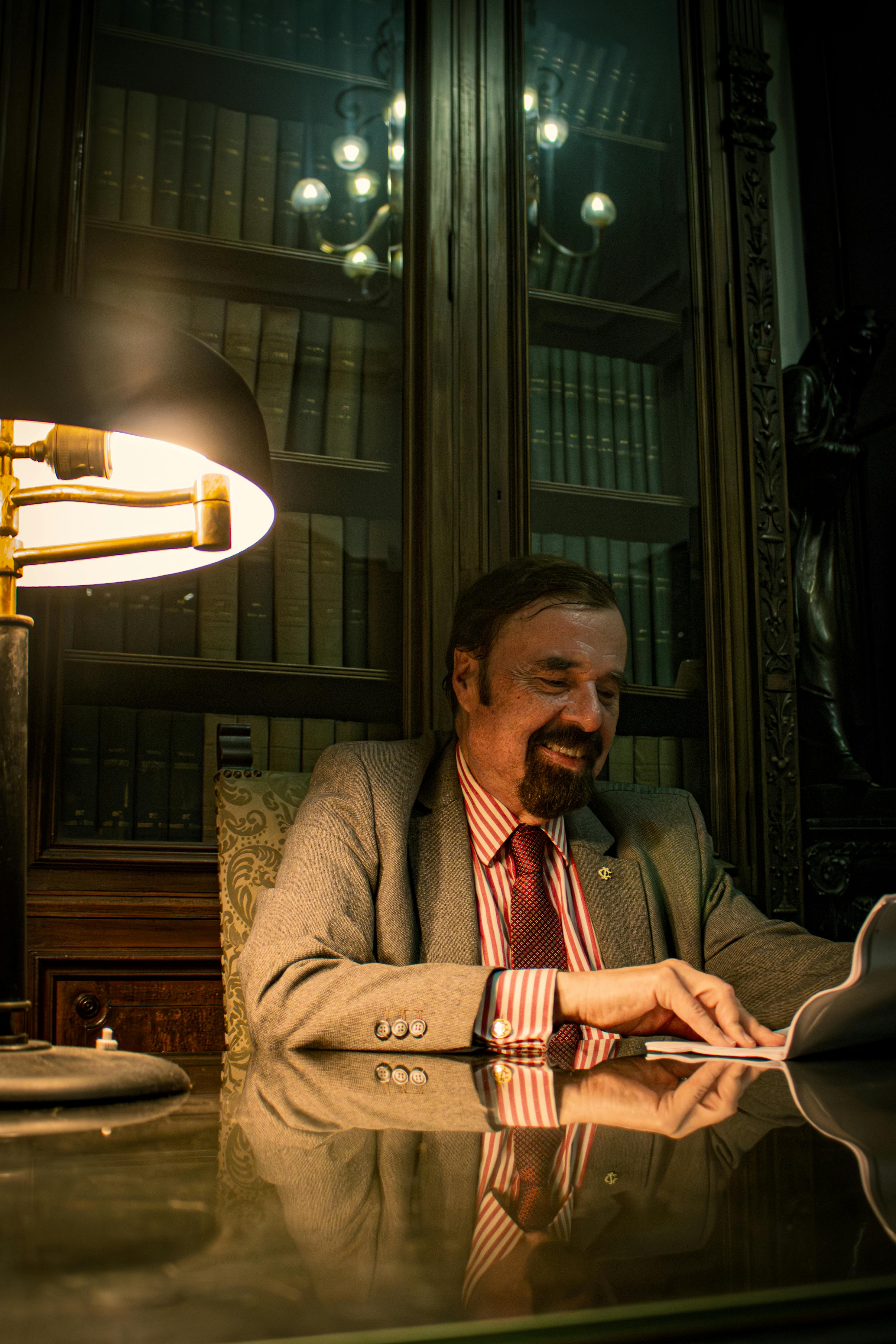 A distinguished man reading in a classic library setting with warm lighting.