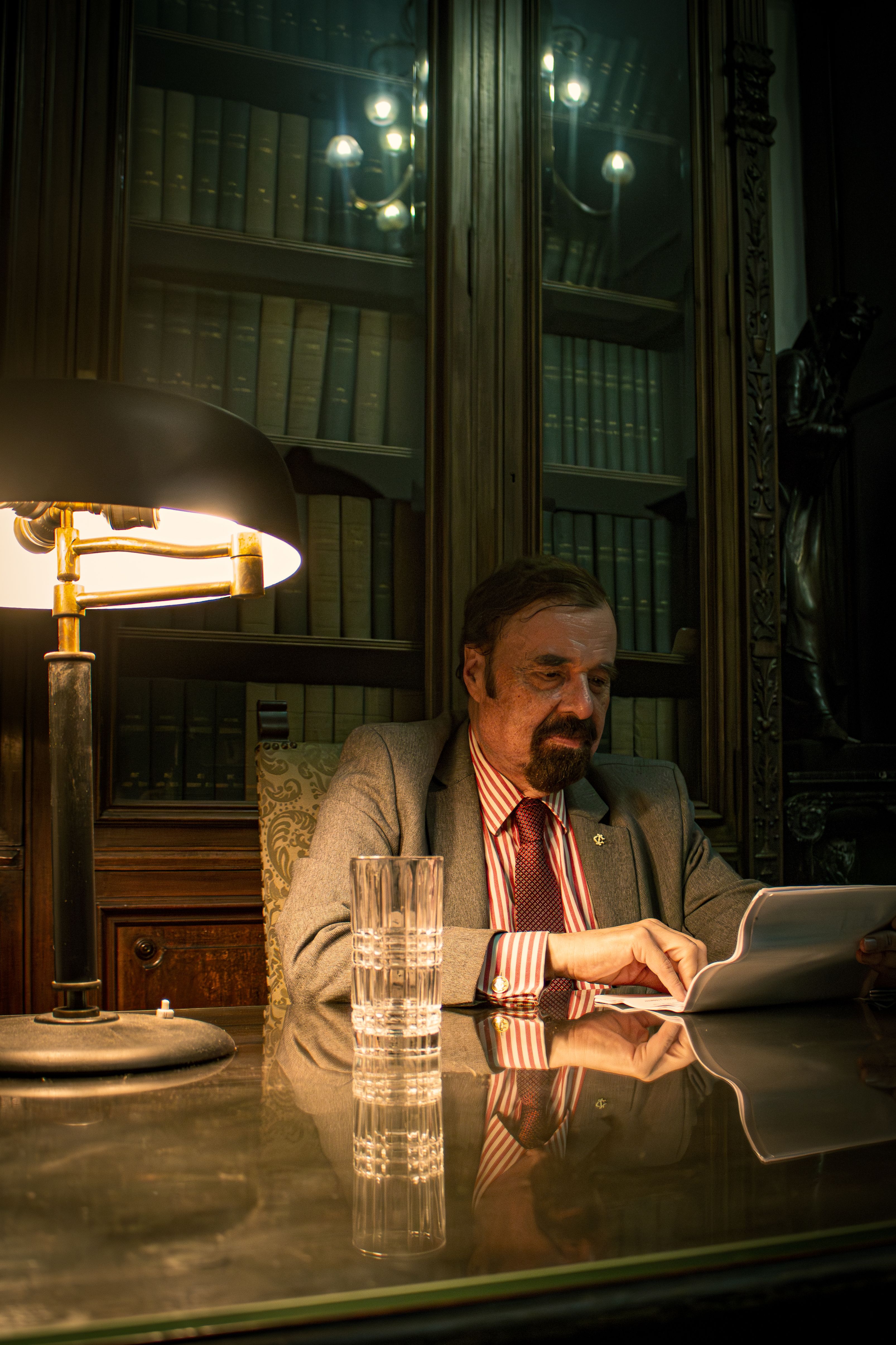 Middle-aged man reading in a dimly lit library with classic decor featuring antique lamps.
