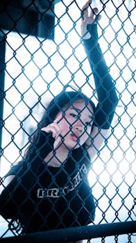 Stylish model poses behind a chain link fence, exuding a dreamy and cinematic vibe in an urban setting.
