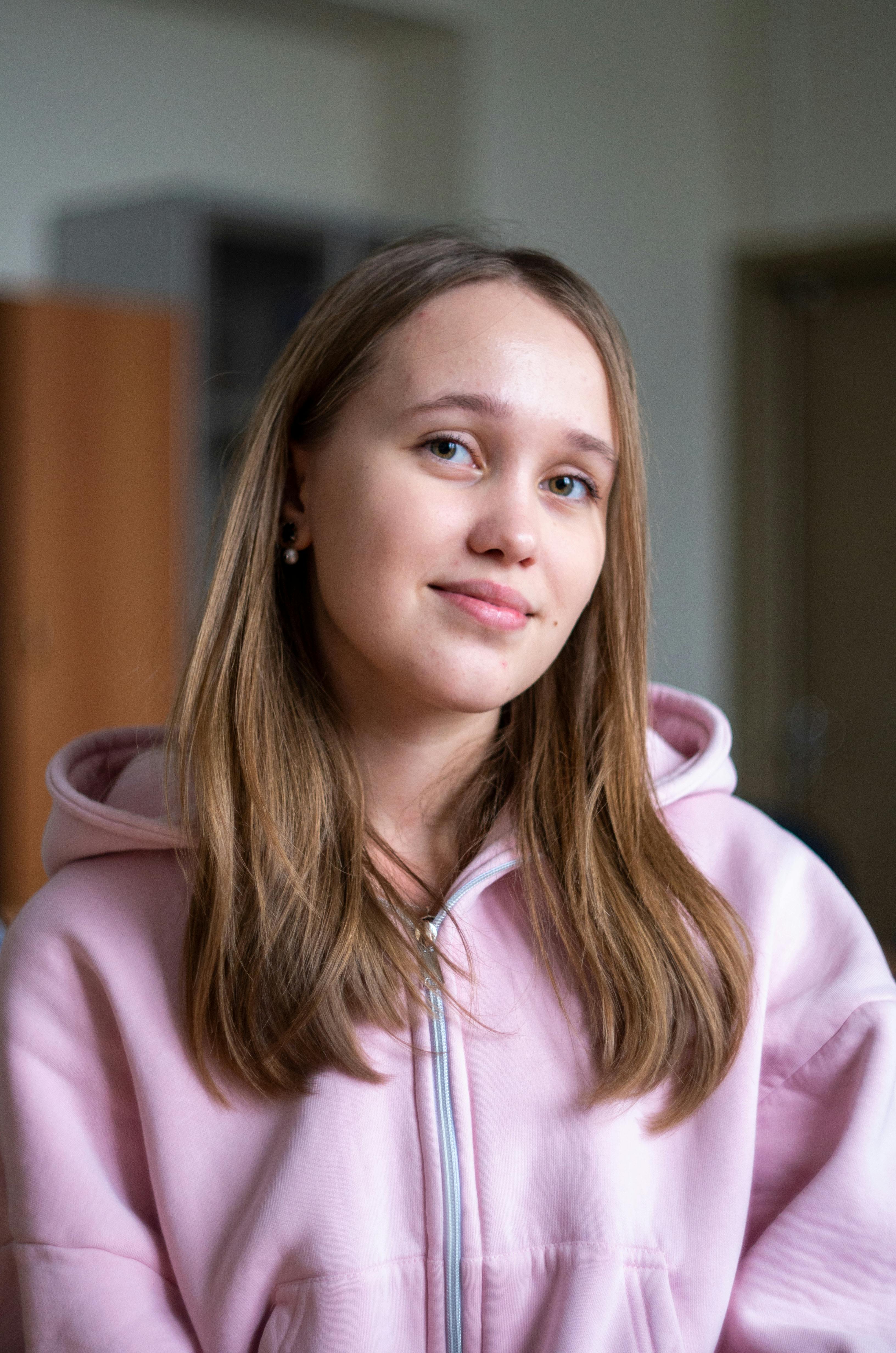 Portrait of a young woman wearing a pink hoodie indoors with a soft smile.