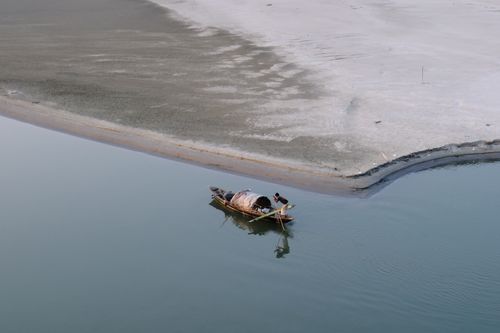 Free A lone fisherman on a boat in a calm river, near a sandy shoreline. Stock Photo