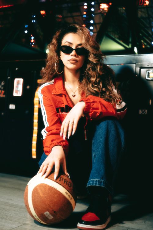 Free Fashionable woman in red jacket posing with basketball in a vibrant arcade setting, exuding a trendy urban vibe. Stock Photo