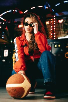 Trendy woman with sunglasses posing with basketball in vibrant arcade setting.