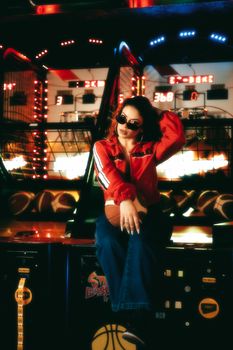 A fashionable woman poses in an arcade wearing sunglasses, creating a vibrant, nostalgic scene.