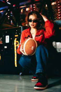 Trendy woman wearing sunglasses and red jacket posing with a football in a lively arcade setting.
