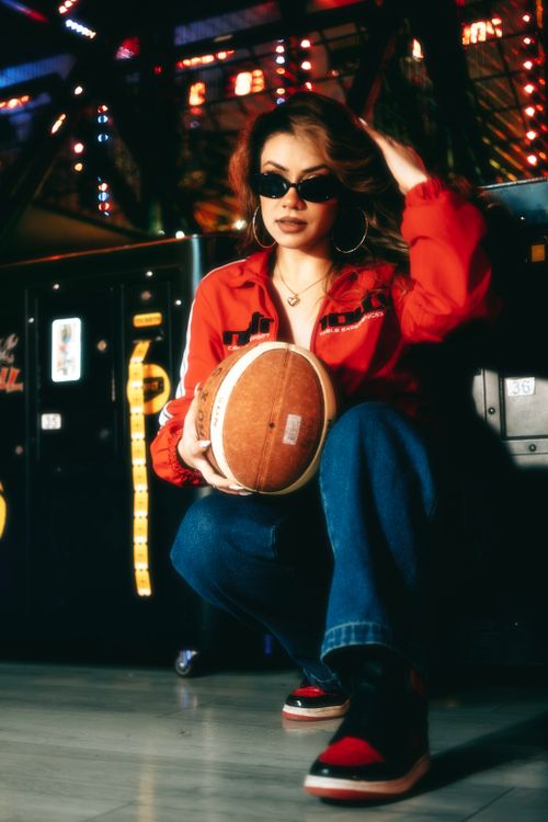 Free Trendy woman wearing sunglasses and red jacket posing with a football in a lively arcade setting. Stock Photo