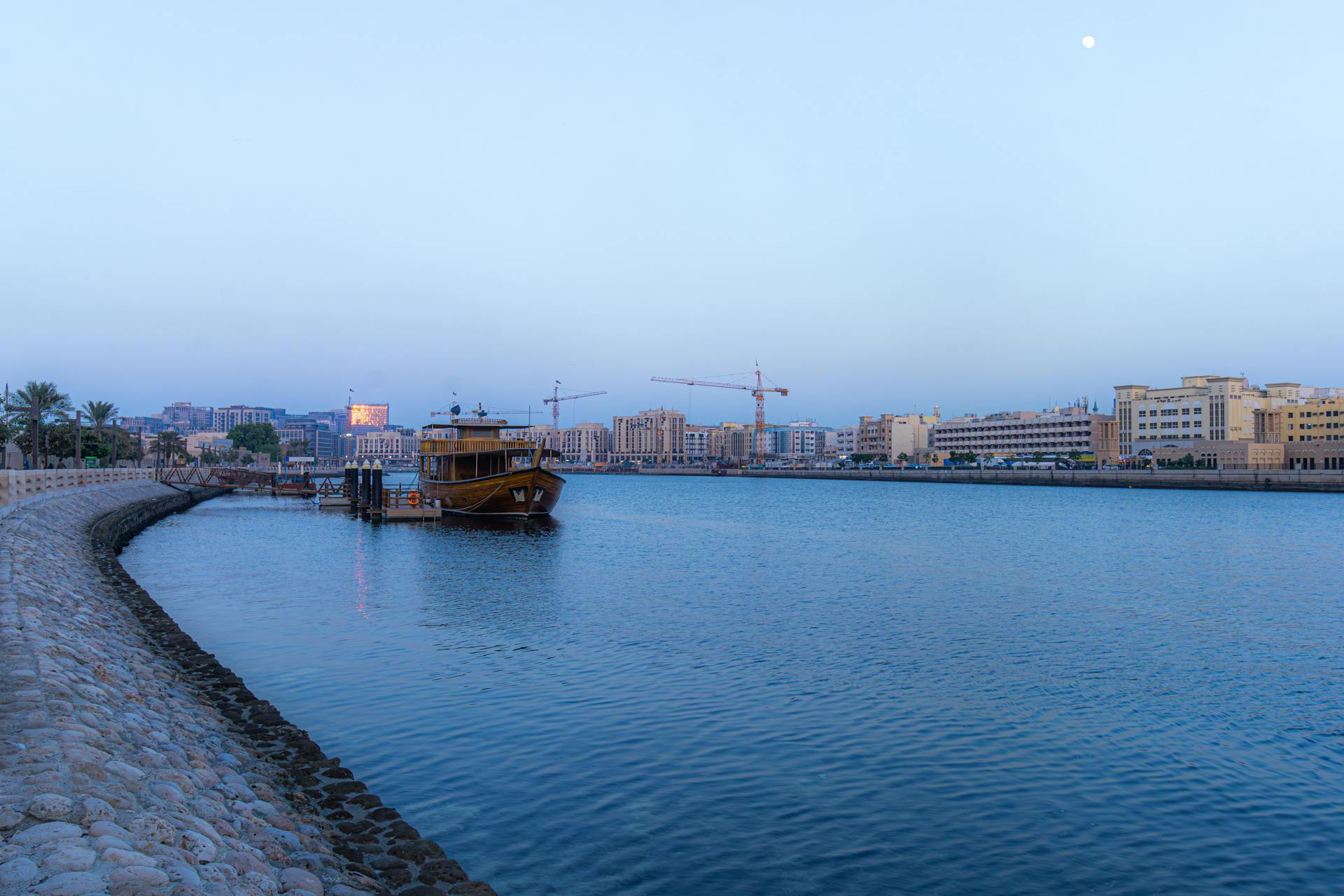 A tranquil view of Dubai Creek with a traditional dhow boat and modern skyline during dusk.