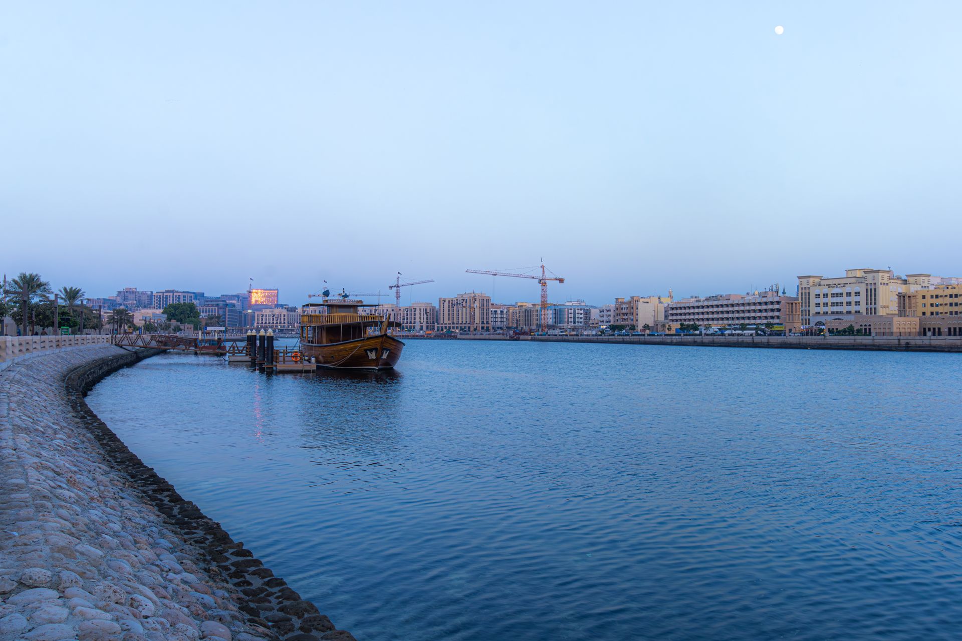 A tranquil view of Dubai Creek with a traditional dhow boat and modern skyline during dusk.