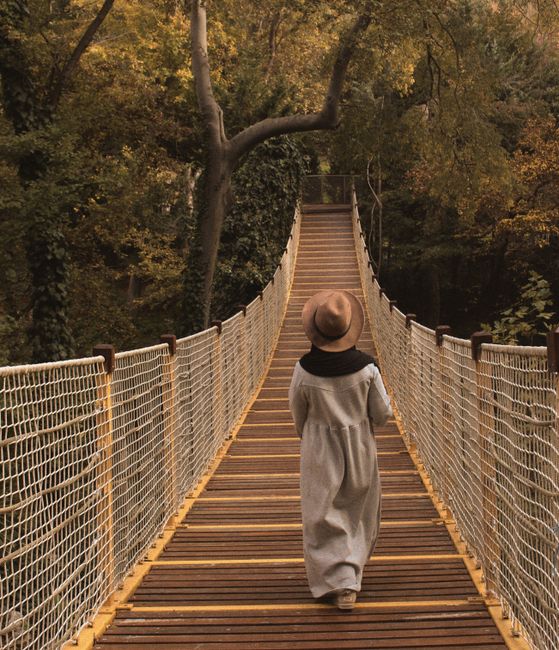 A woman in autumn attire walks across a wooden suspension bridge in a forest setting.