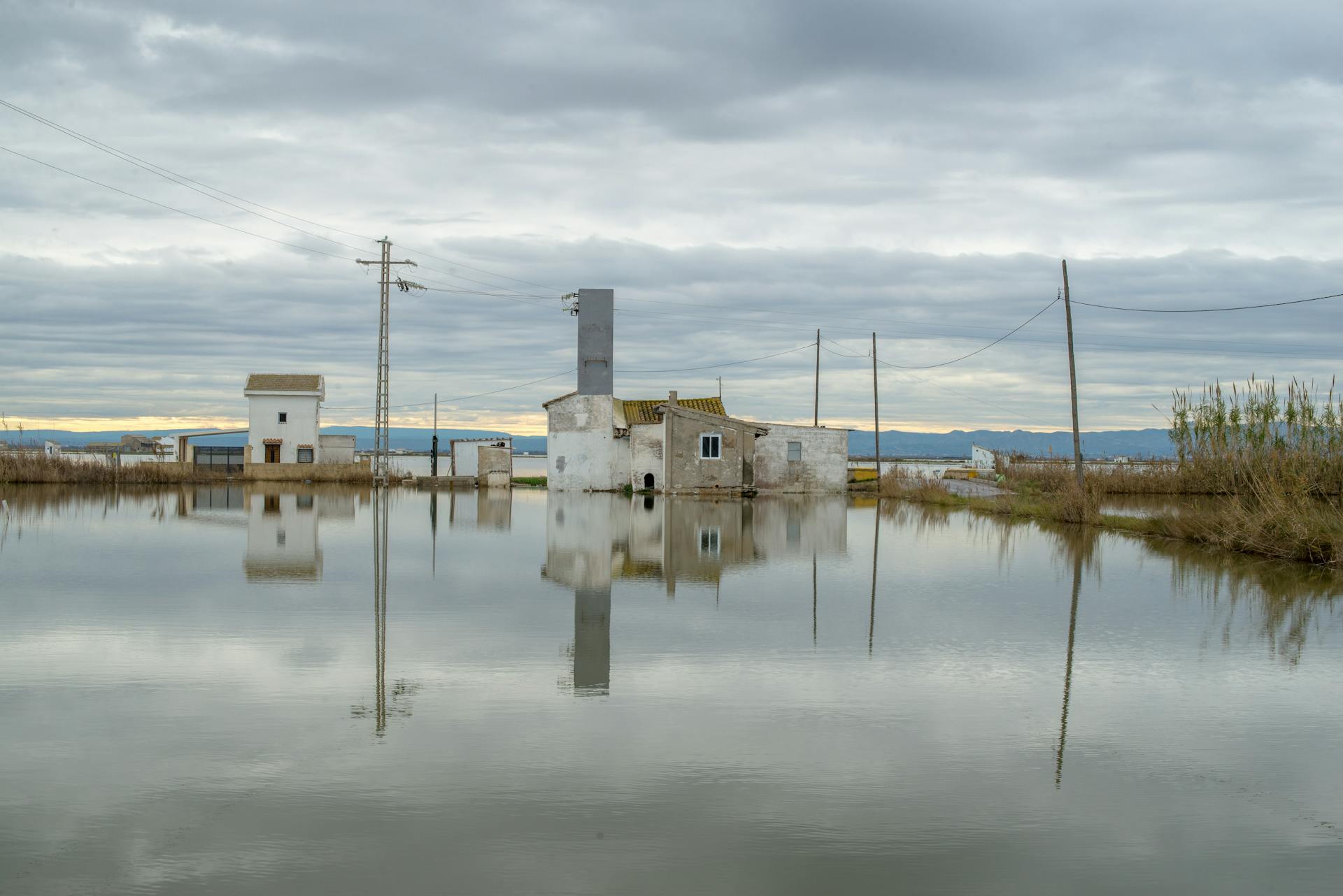 A serene rural scene with buildings reflecting on a flooded landscape under cloudy skies.