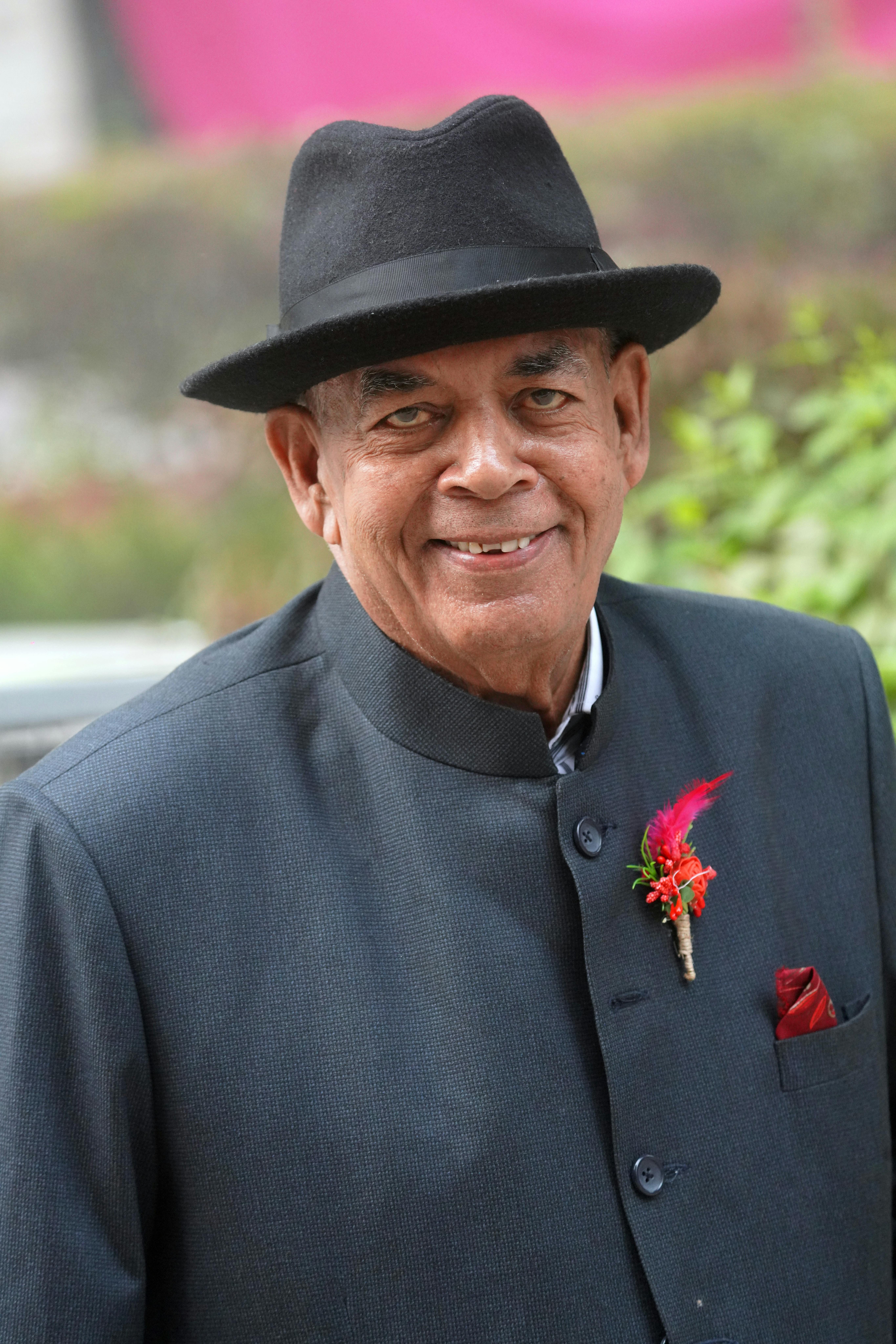 Portrait of an elderly man wearing a formal suit and fedora, smiling outdoors.