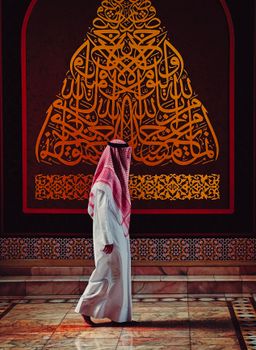 A man in traditional Middle Eastern attire walks past intricate Arabic calligraphy on a tiled floor indoors.