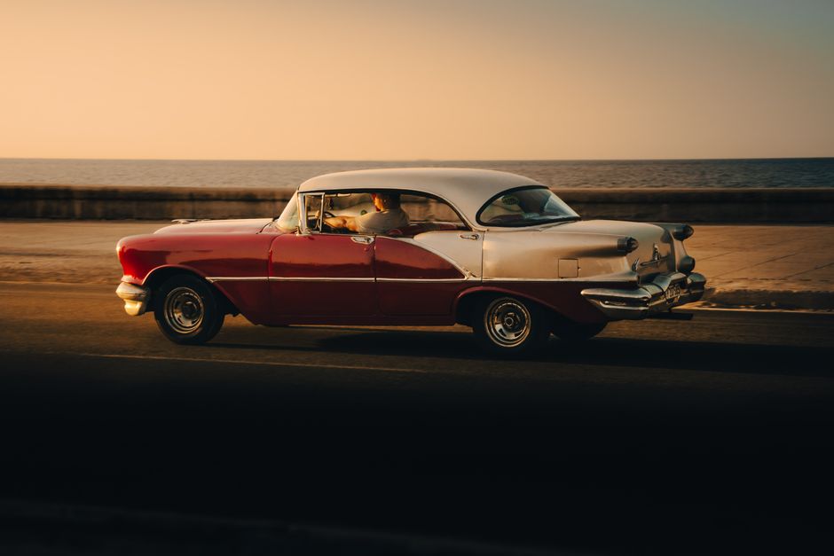 A classic vintage car with two-tone paint driving by the seaside during sunset.