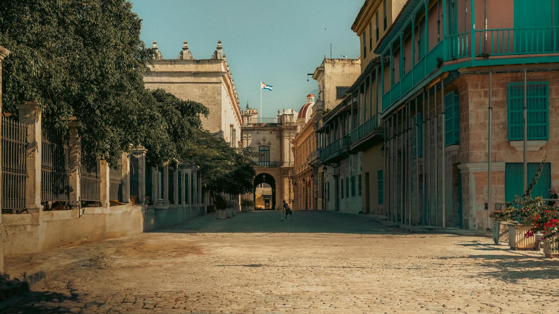 Sunlit historic street in Havana with colonial buildings and Cuban flag.