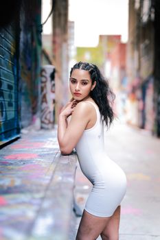 A fashionable woman in a white dress poses in a vibrant graffiti-covered alleyway.