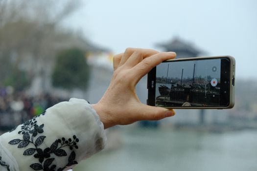 A hand holds a smartphone capturing a serene Chinese scene, blending technology and tradition.