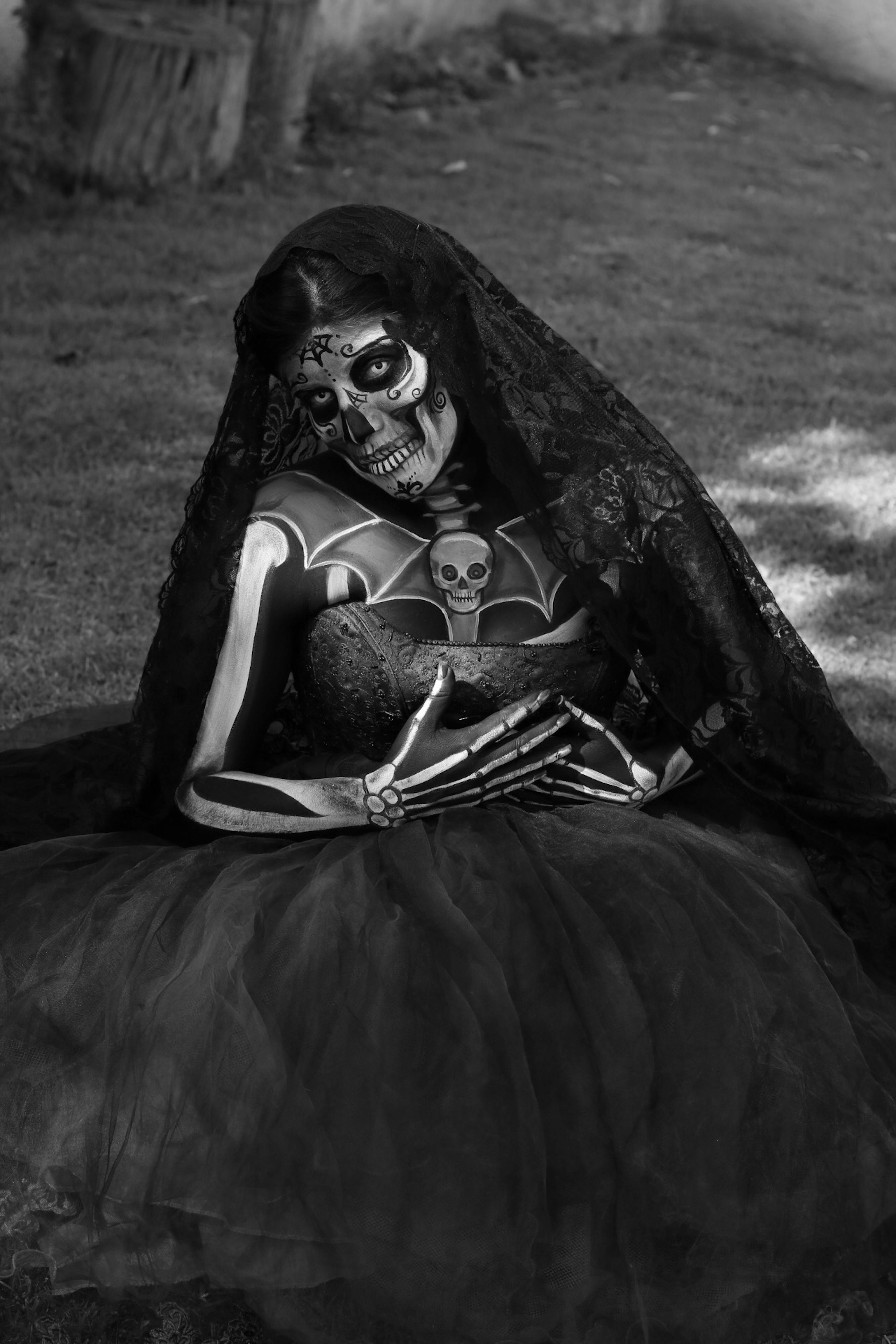 Ücretsiz Açık havada çimenlerin üzerinde oturan kafatası makyajlı ve gotik kıyafetli bir kadının siyah beyaz portresi. Stok Fotoğraflar