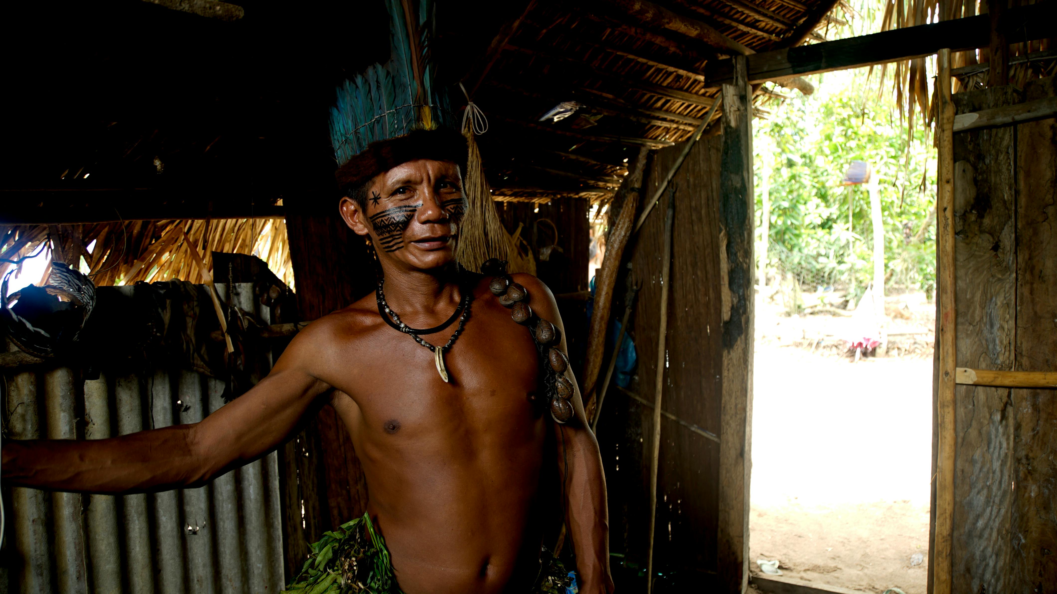Portrait of an indigenous man in traditional attire inside a rustic dwelling in Amazonas, Brazil.