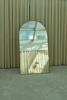 A stylish mirror reflects a sunlit interior with curtains and modern architecture.