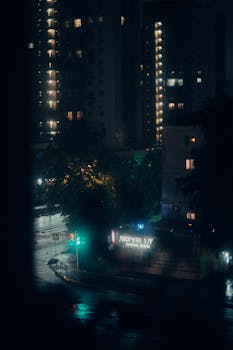 A cinematic view of a rainy city street at night with illuminated buildings.