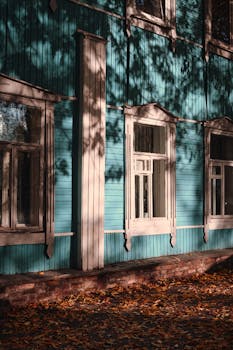 Sunlit wooden cottage wall with autumn leaves on the ground, evoking cottagecore vibes.