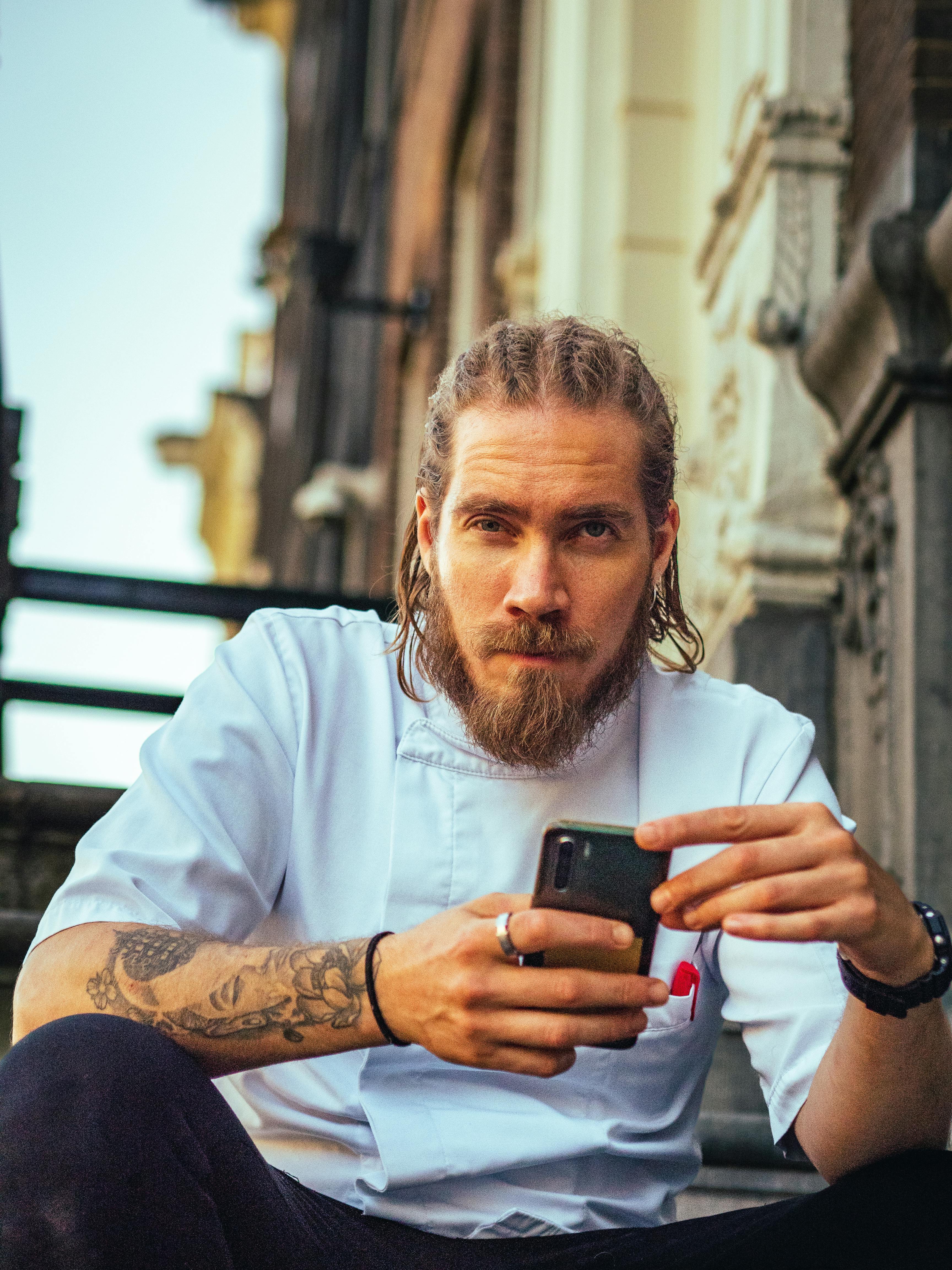 Casual portrait of a bearded man sitting on steps in Amsterdam, holding a smartphone.