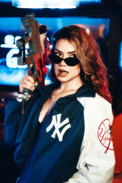 Free Confident woman in sunglasses holding a toy gun in a vibrant neon arcade setting. Stock Photo