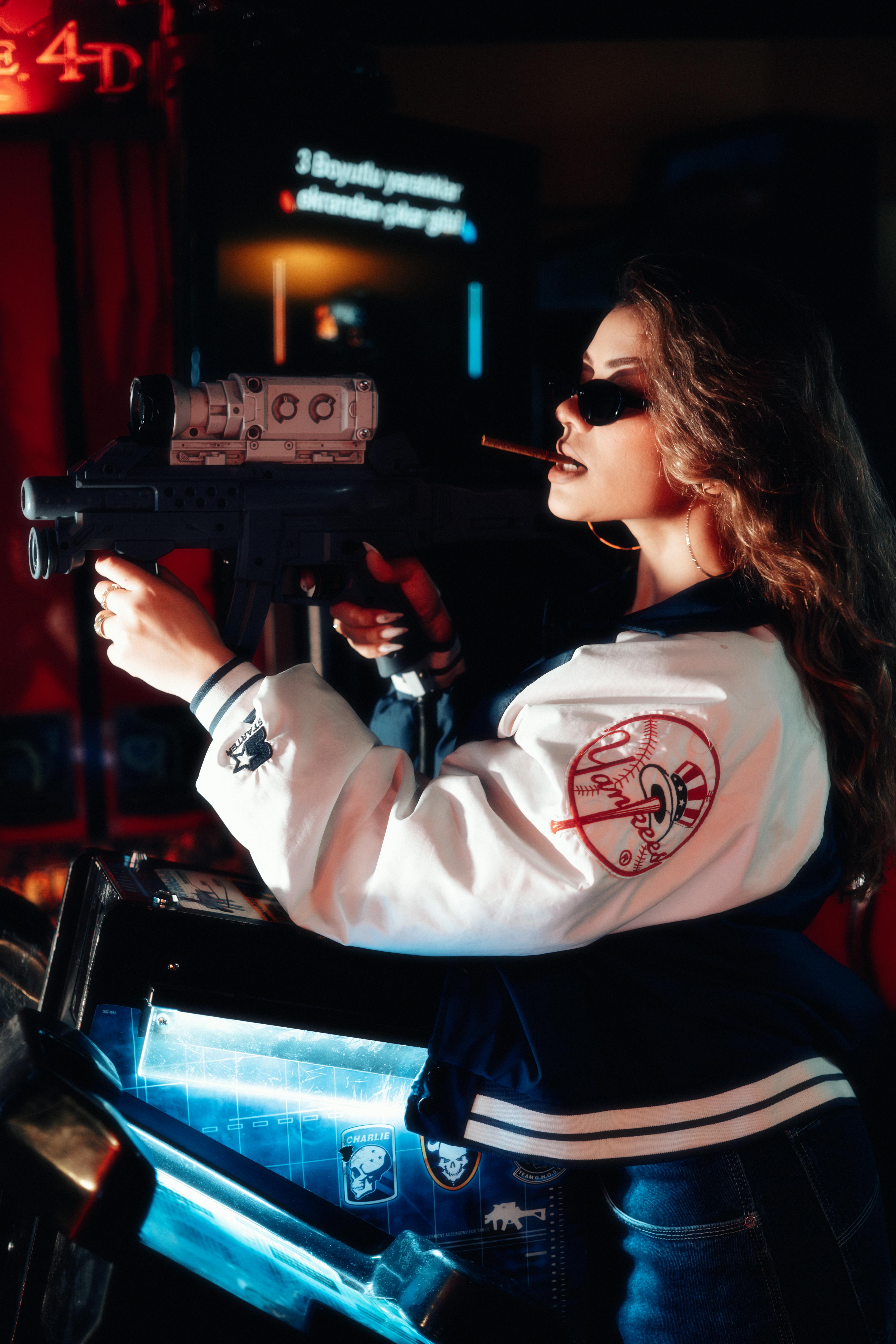 Young woman in varsity jacket plays arcade shooter game with sunglasses and toy gun.