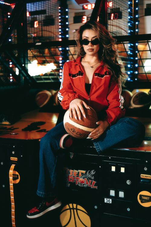 Free Stylish woman in red jacket poses with basketball in a vibrant arcade setting. Stock Photo