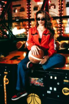 Stylish woman in red jacket posing with basketball in a vibrant arcade setting.