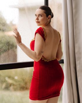 A woman in a red backless dress gazes out a rain-speckled window indoors.