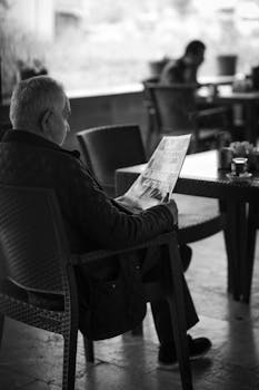 Black and white photo of an elderly man reading a newspaper in an outdoor café.
