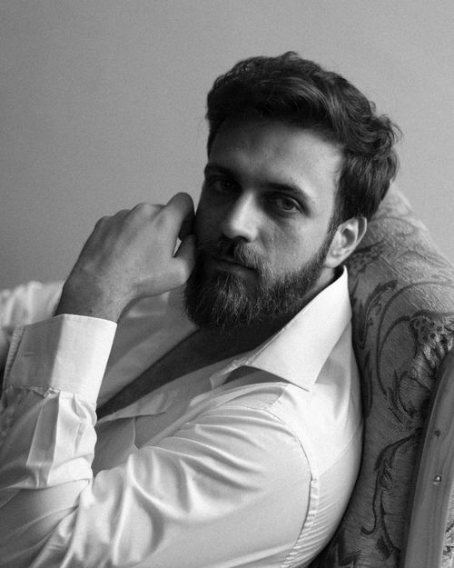 Free Black and white portrait of a bearded man in a white shirt, reclining on a couch, gazing intently at the camera. Stock Photo