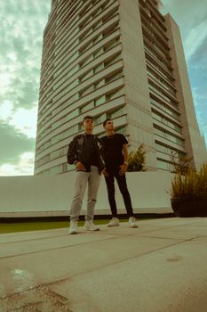 Two men pose confidently in front of a modern high-rise building under a cloudy sky.