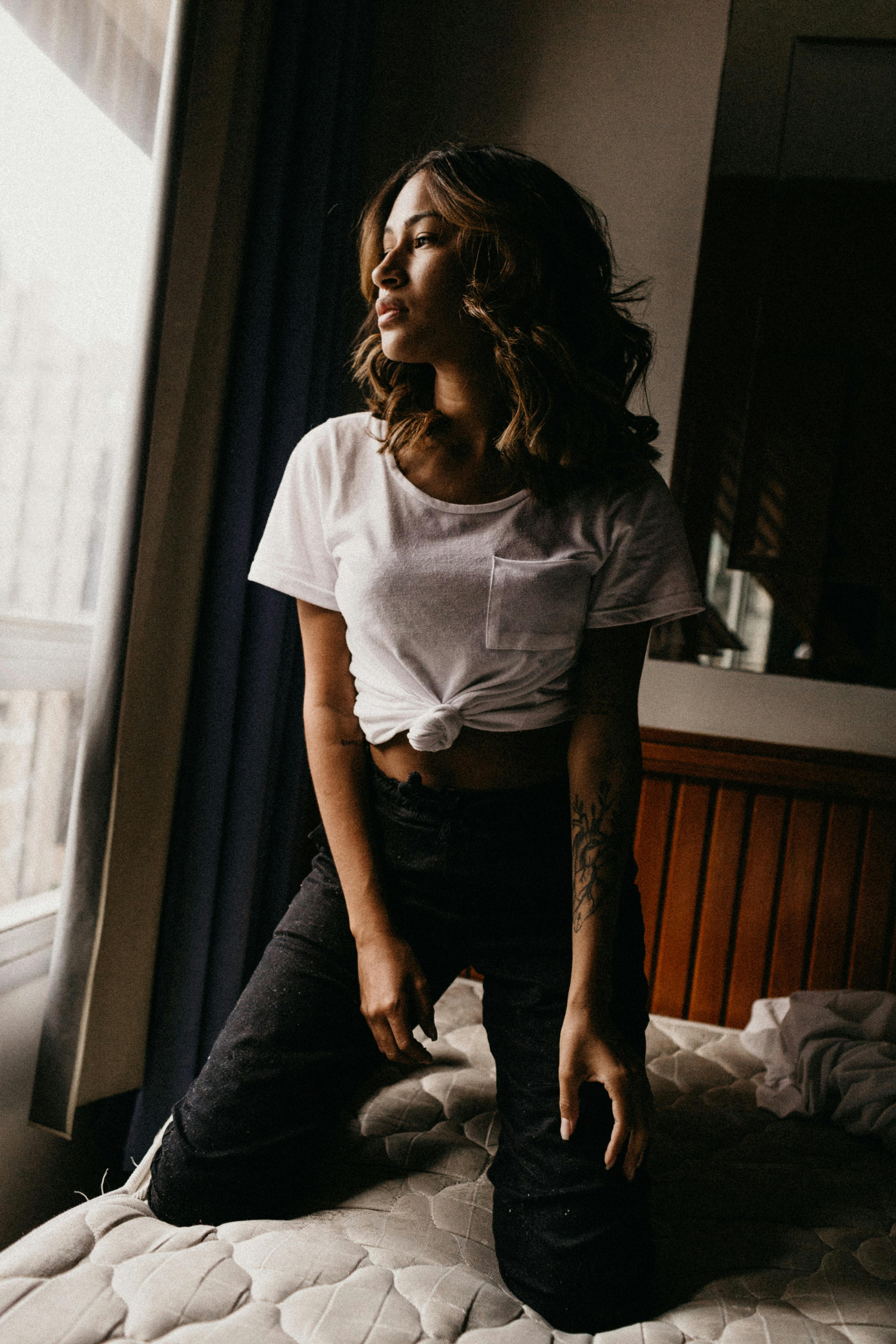 Young woman in casual outfit posing elegantly on bed in sunlit bedroom.