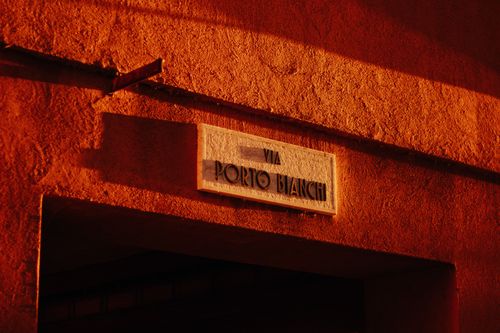 Free A vintage street sign 'Via Porto Bianchi' illuminated by warm sunset lighting, creating a dramatic effect. Stock Photo